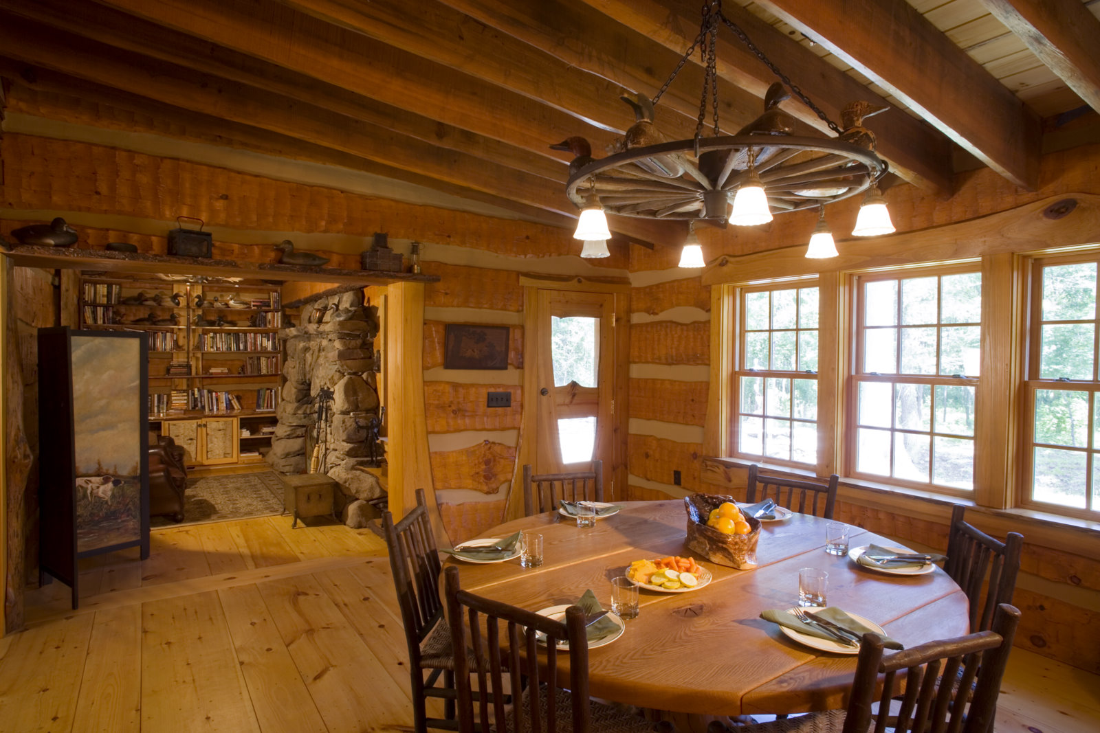 Dining area with wagon-wheel chandelier, chinked log walls, and view into a stone-fireplace library