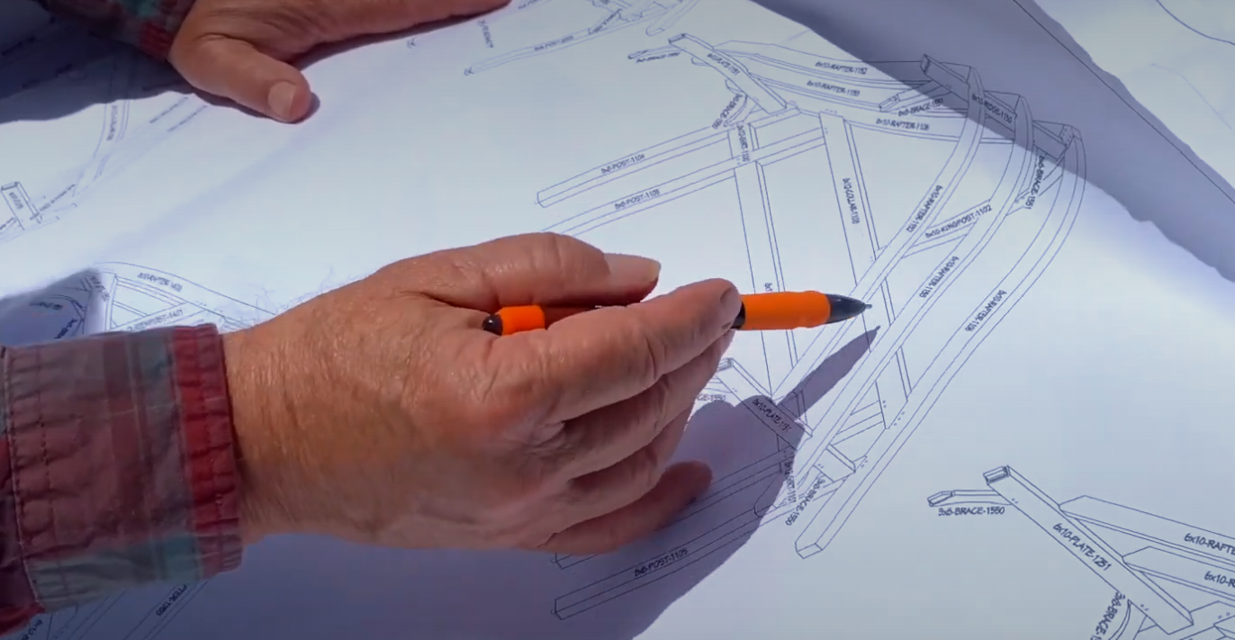 Hands reviewing timber frame shop drawings with labeled posts, rafters, and braces