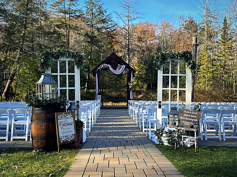 Timber ceremony arbor dressed in greenery at the creekside site, autumn woods behind
